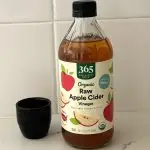 A bottle of Whole Foods 365 raw apple cider vinegar next to a tiny black vase on a marble kitchen counter.