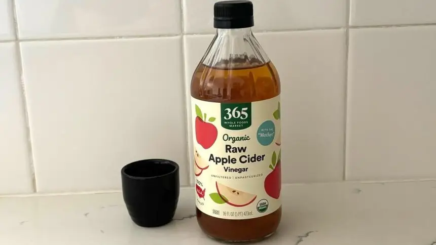 A bottle of Whole Foods 365 raw apple cider vinegar next to a tiny black vase on a marble kitchen counter.