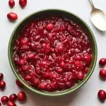 A serving dish containing cranberry sauce. A spoon rests at the top of the bowl and there are whole cranberries scattered around the bowl.