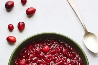 A serving dish containing cranberry sauce. A spoon rests at the top of the bowl and there are whole cranberries scattered around the bowl.