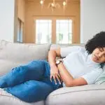 Woman laying on gray couch holding stomach with eyes closed