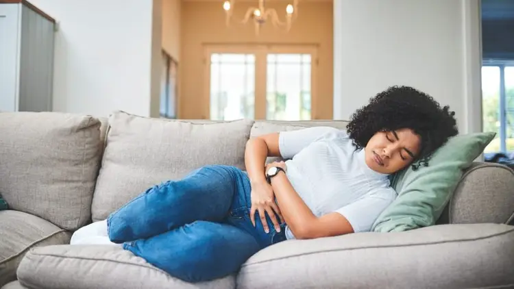 Woman laying on gray couch holding stomach with eyes closed