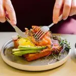 A man&apos;s hands cutting a salmon and avocado flatbread with a knife and fork.