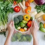 Close-up of hands placing fresh vegetables and fruits into an eco-mesh bag on a white table.