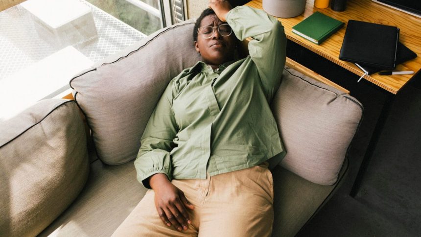 A woman laying on a light gray sofa next to a desk with her hand on her head.