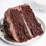 A slice of chocolate cake with chocolate buttercream frosting on a white plate with a metal fork to the side.