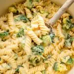 Rotini pasta, broccoli and spinach in a pot coated in a cheese sauce. A wooden spoon is scooping up some of the pasta.