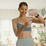 A person with pulled-back dark brown hair and a medium-blue sports bra pouring a smoothie into a glass.