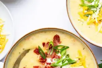 A bowl of potato soup with a silver spoon in it. The soup is topped with bacon bits, green onion, shredded cheese and sour cream.