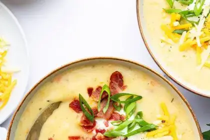 A bowl of potato soup with a silver spoon in it. The soup is topped with bacon bits, green onion, shredded cheese and sour cream.