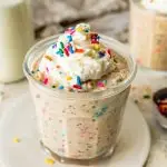 A glass jar filled with birthday cake overnight oats, topped with whipped cream and colorful sprinkles, with a bottle of milk and another jar of oats in the background.