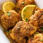 Six baked Greek chicken thighs in a white baking dish. The chicken is topped with fresh parsley and lemon slices.