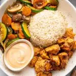 A plate featuring hibachi chicken, brown rice, and sautéed vegetables with a side of creamy dipping sauce.