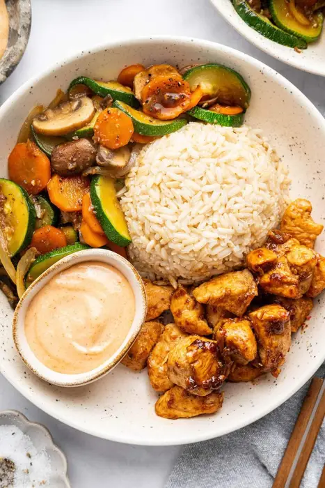 A plate featuring hibachi chicken, brown rice, and sautéed vegetables with a side of creamy dipping sauce.