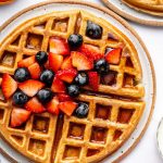 Golden brown protein waffles on a plate, topped with fresh strawberries and blueberries, drizzled with syrup, and served with a side of fruit.