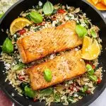 Two salmon filets in a skillet served over a bed of orzo combined with roasted red peppers, chopped dates, feta and swiss chard. It