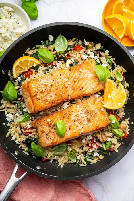 Two salmon filets in a skillet served over a bed of orzo combined with roasted red peppers, chopped dates, feta and swiss chard. It