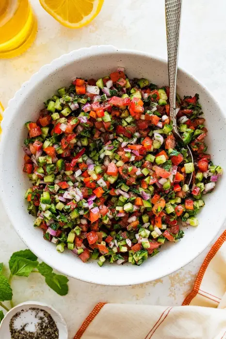 A Shirazi Salad in a white bowl served with a silver spoon. There is a lemon half and a bottle of olive olive next to the bowl.