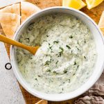 A white bowl of tzatziki topped with fresh dill. There is a wooden spoon in the bowl and the bowl is surrounded by pita bread and lemon slices.