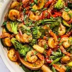 Shrimp stir-fry with shrimp, broccoli, red bell peppers, snap peas, zucchini, and onions coated in sauce. The dish is garnished with sesame seeds and chopped green onions, with a wooden spoon in the pan.