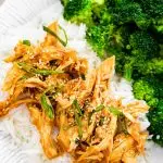 Slow cooker teriyaki chicken served on a plate with white rice and broccoli. The chicken is topped with sesame seeds and green onions.