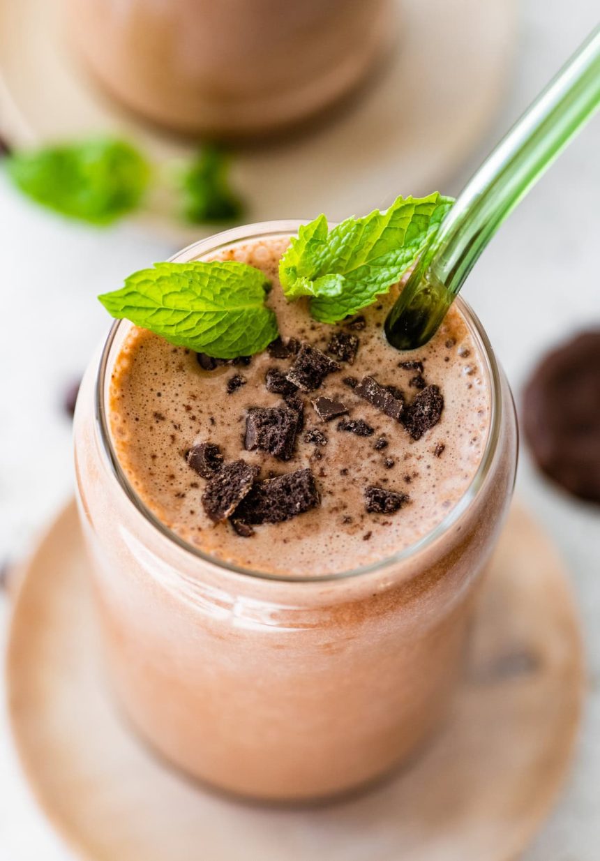 One mint chocolate protein shake in a glass topped with crushed Thin Mint cookies and fresh mint. There is a green straw in the glass.