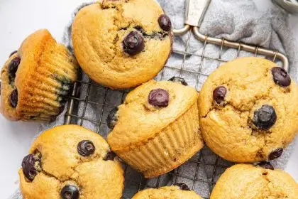 12 blueberry cottage cheese muffins spread out over a wire cooling rack. There is a light blue towel under the cooling rack and one of the muffins has a bite taken out of it.