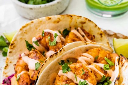 Three shrimp tacos in flour tortillas folded up on a serving platter. The tacos are topped with a mango cabbage slaw and a drizzle of spicy crema.