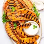 Grilled sweet potatoes on a serving platter topped with fresh parsley and served with a side of garlic aioli. There is one sweet potato dipped in the aioli.