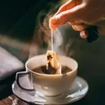 A person dunking a tea bag into a cup of tea a steaming cup on a wooden surface