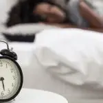Close-up photo of an alarm clock and a woman sleeping in the background.